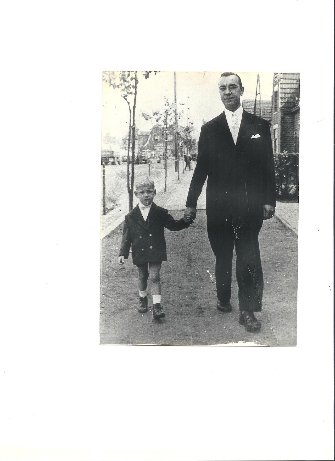 A man in a suit walks down the sidewalk holding hands with a young boy in a blazer.