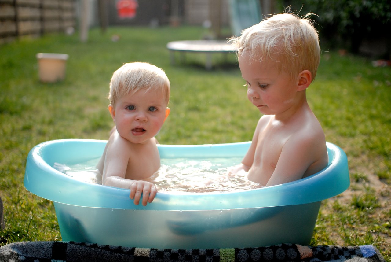 Two young children sit in a small plastic tub filled with water, enjoying an outdoor bath on the grass.