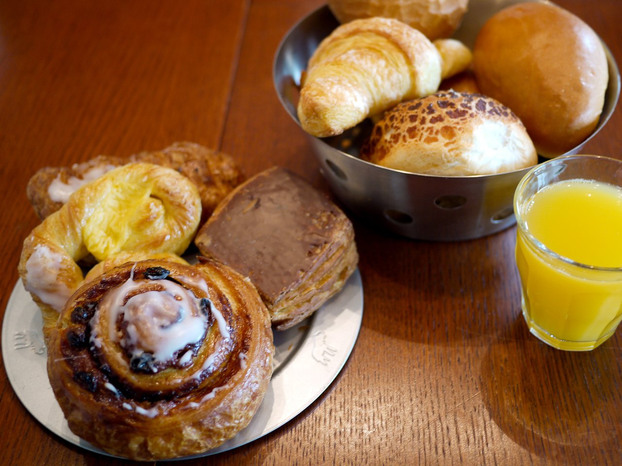 A plate of pastries, a basket of bread, and a glass of orange juice on a wooden table.