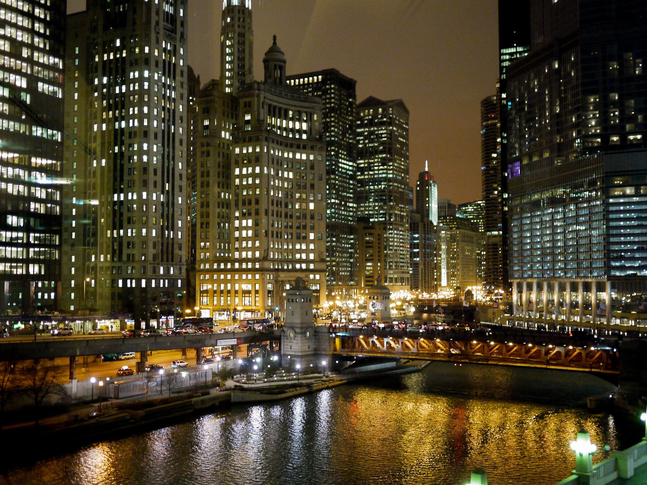 Chicago skyline at night with illuminated skyscrapers, a lit bridge, and city lights reflecting on the river.