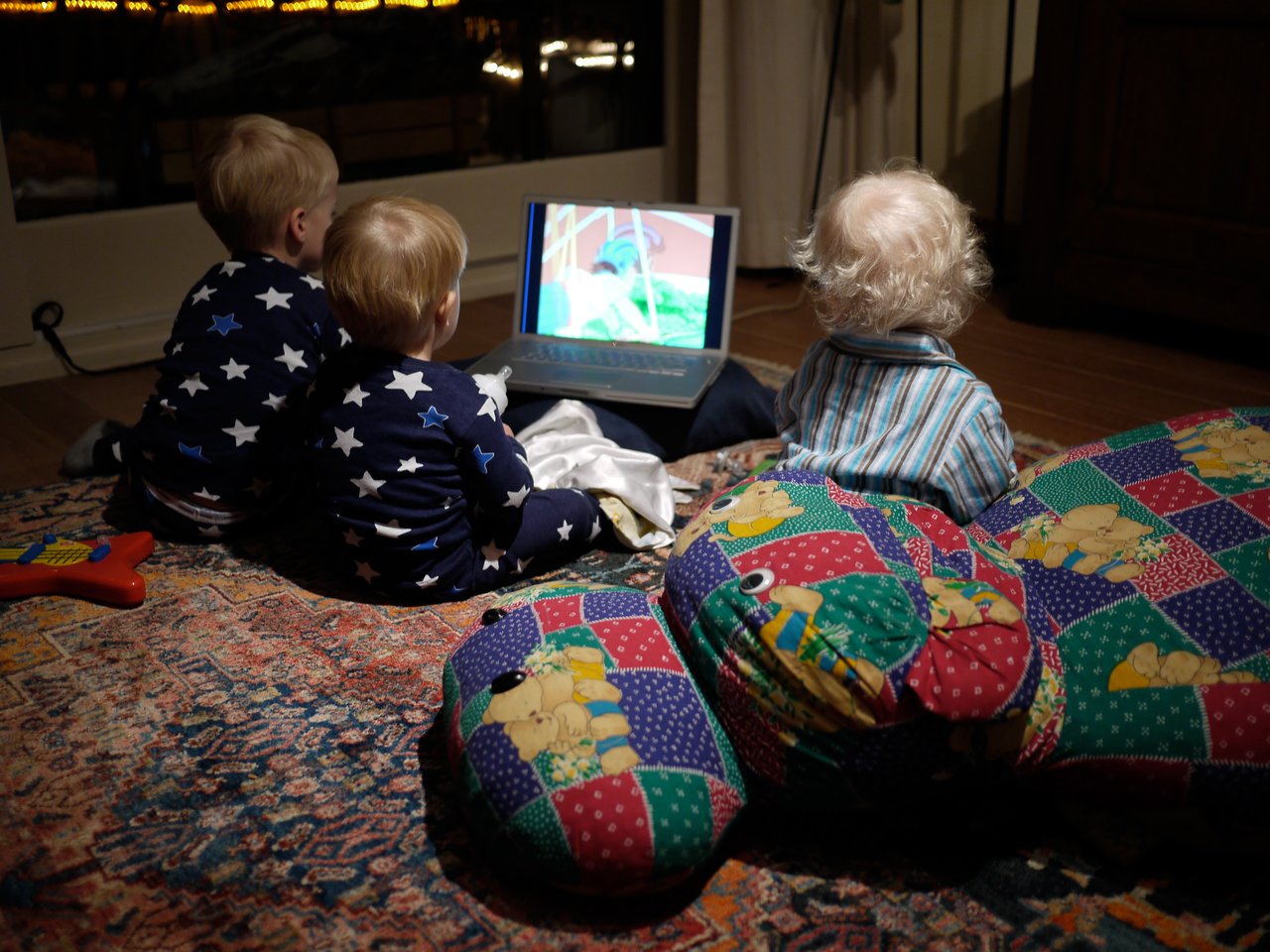Three young children in pajamas sit on the floor, watching a laptop screen in a dimly lit room.