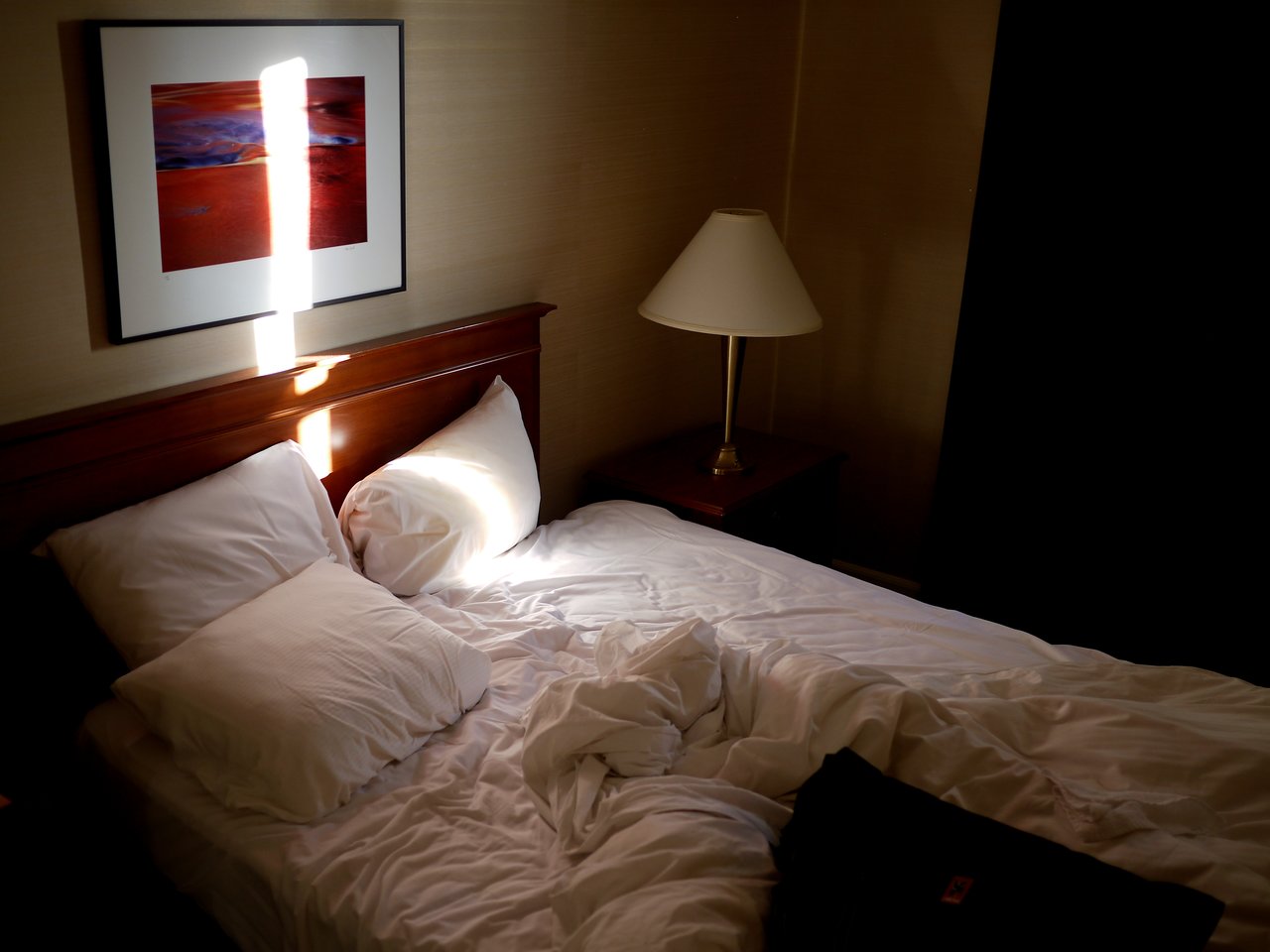 An unmade hotel bed with two pillows, a bedside lamp, and sunlight creating a bright stripe on the wall.