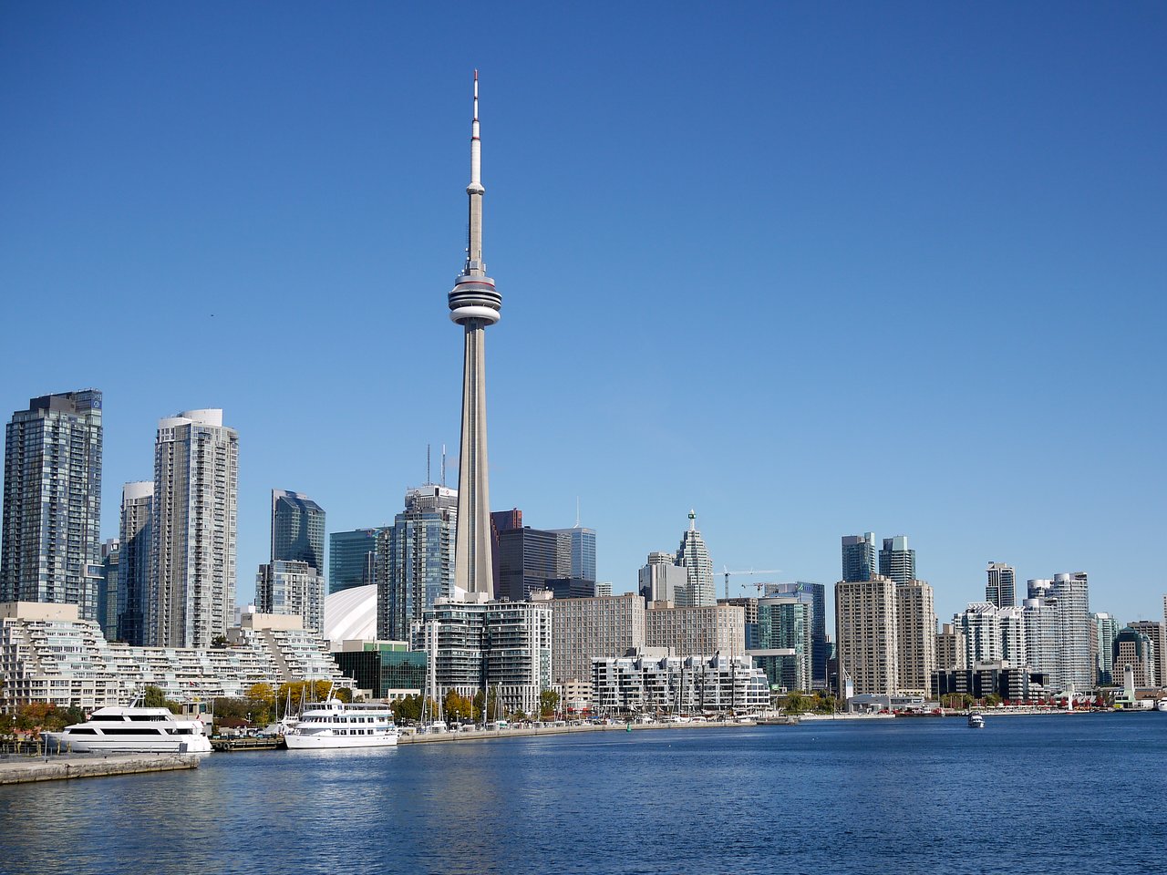 Toronto skyline featuring the CN Tower, high-rise buildings, and boats on the waterfront under a clear blue sky.