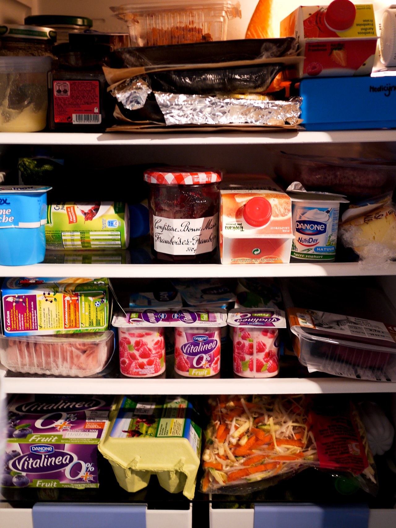 A refrigerator filled with yogurt, jam, juice, eggs, vegetables, and packaged meat, neatly arranged on multiple shelves.