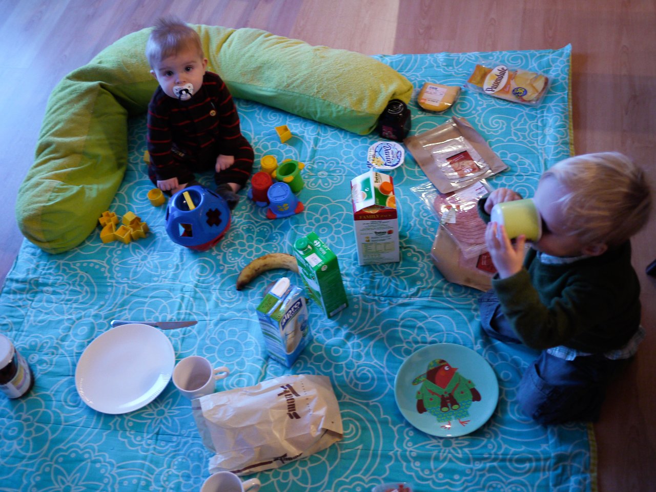Two young children sit on a blanket indoors, surrounded by breakfast food and toys.