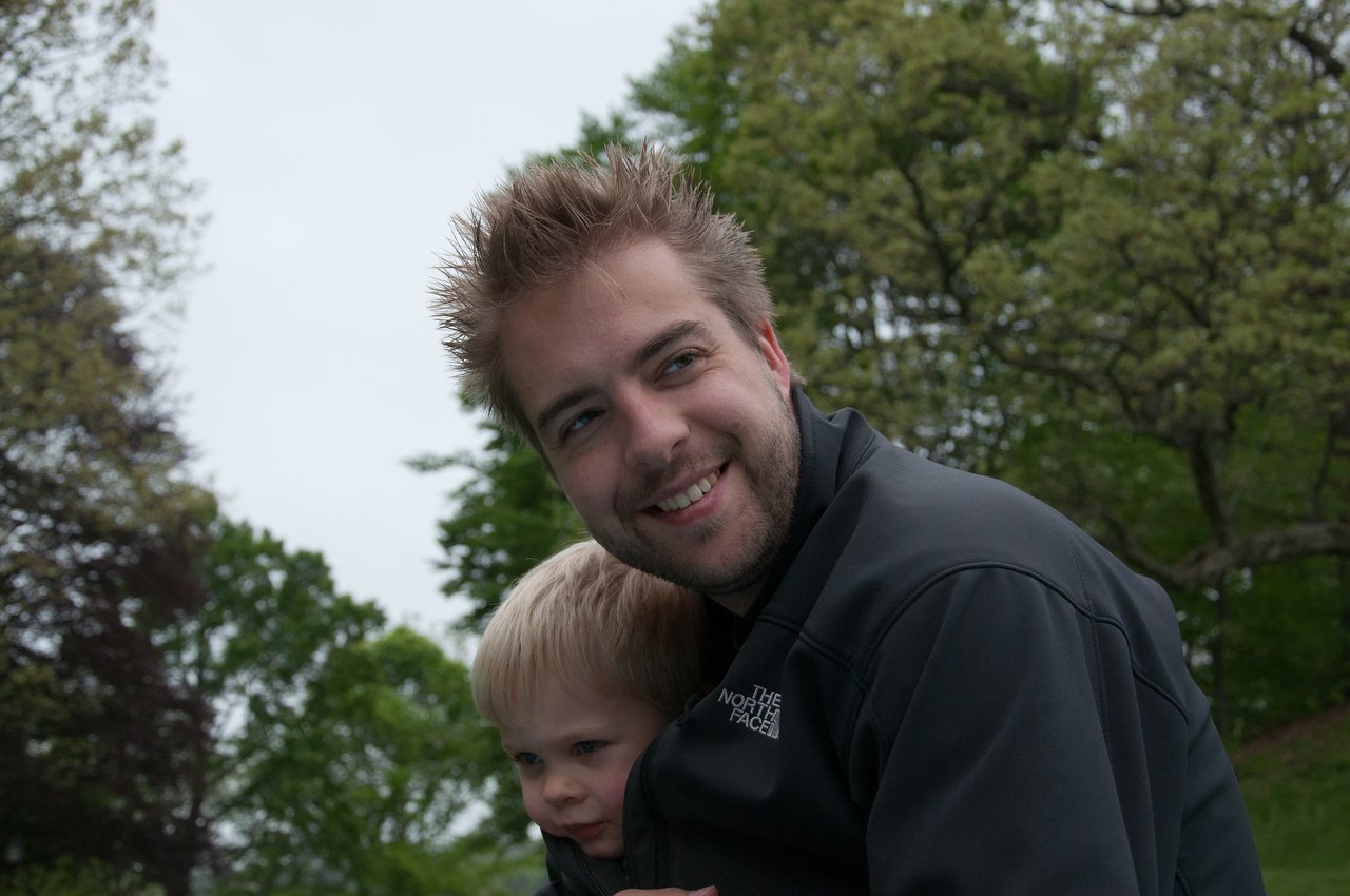 A man smiles while holding a young child close to him outdoors with trees in the background.
