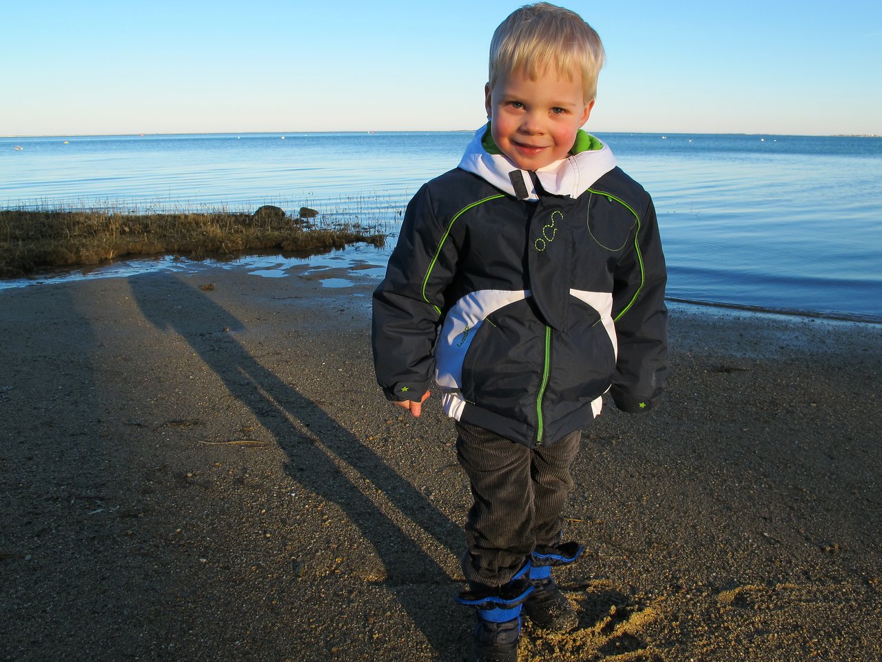 A young child in a jacket and boots stands on a sandy beach, smiling near the water.