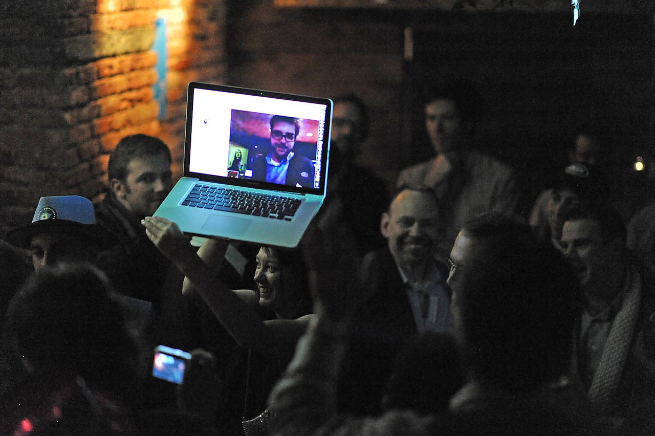 A woman holds up a laptop showing a video call while a crowd celebrates at a Drupal release party.