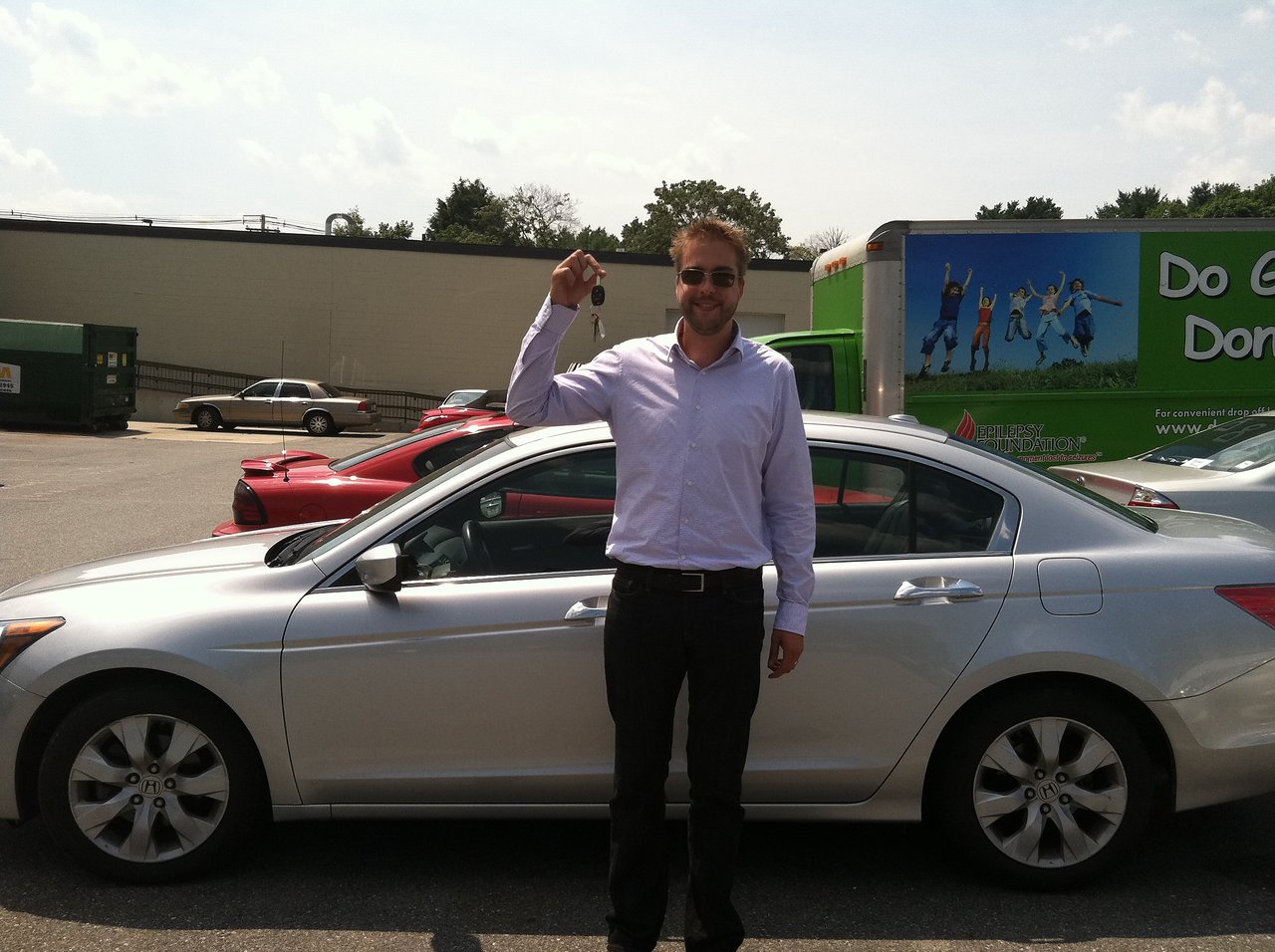 A man in a white shirt stands in front of a silver car, holding up a car key.