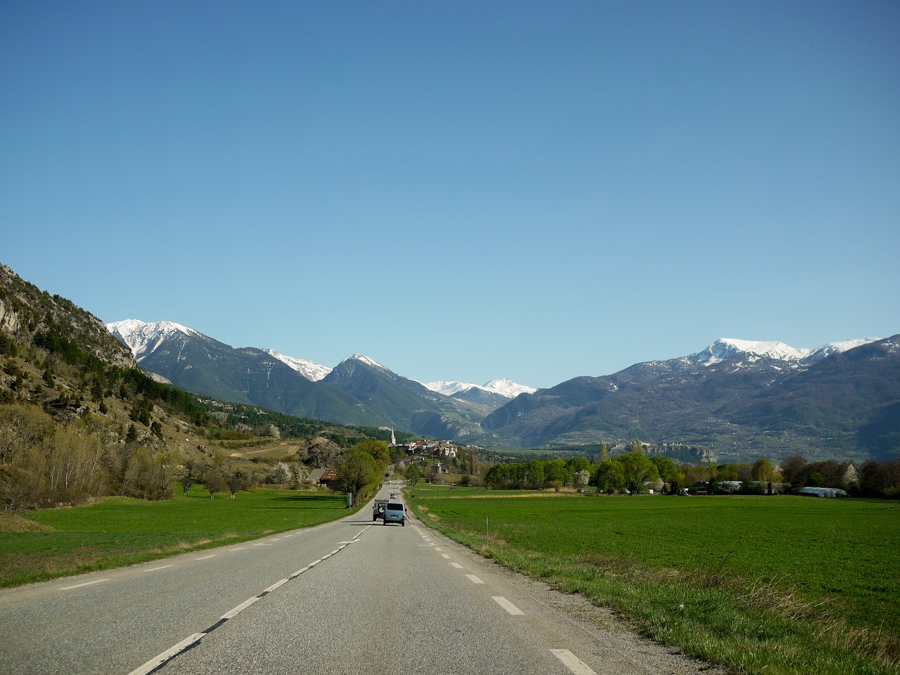 A car drives on a two-lane road toward snow-capped mountains in the French Alps under a clear sky.