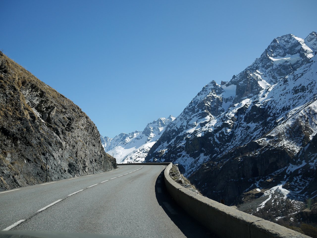 A winding mountain road in the French Alps, surrounded by rocky cliffs and snow-covered peaks under a clear sky.