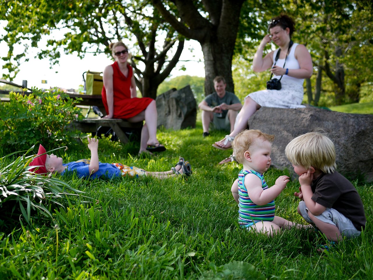 Two young children sit facing each other in the grass, while others relax nearby in a park-like setting.