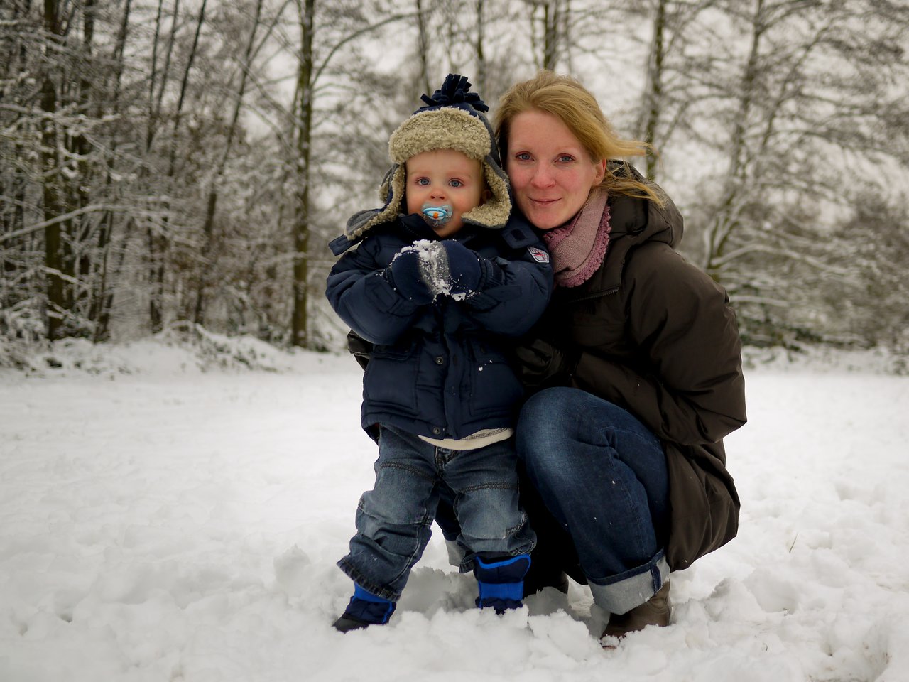 A woman and a young child in winter clothing pose together in the snow, with trees in the background.