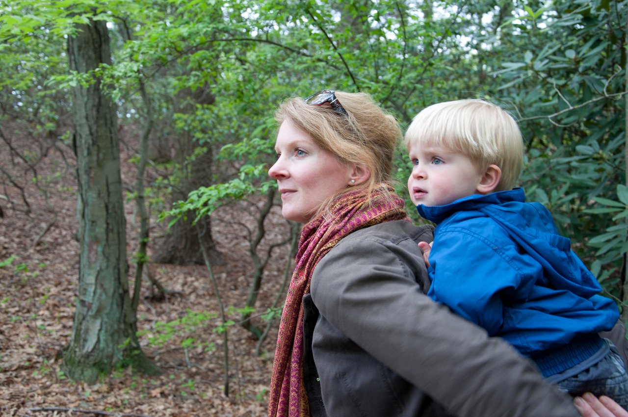 A woman carries a young child on her back while walking through a forest.