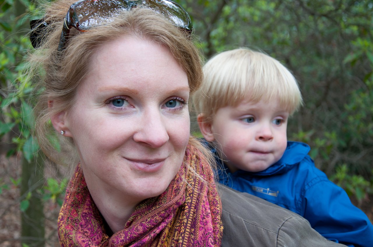 A woman smiles at the camera while a young child in a blue jacket sits on her back, looking away.