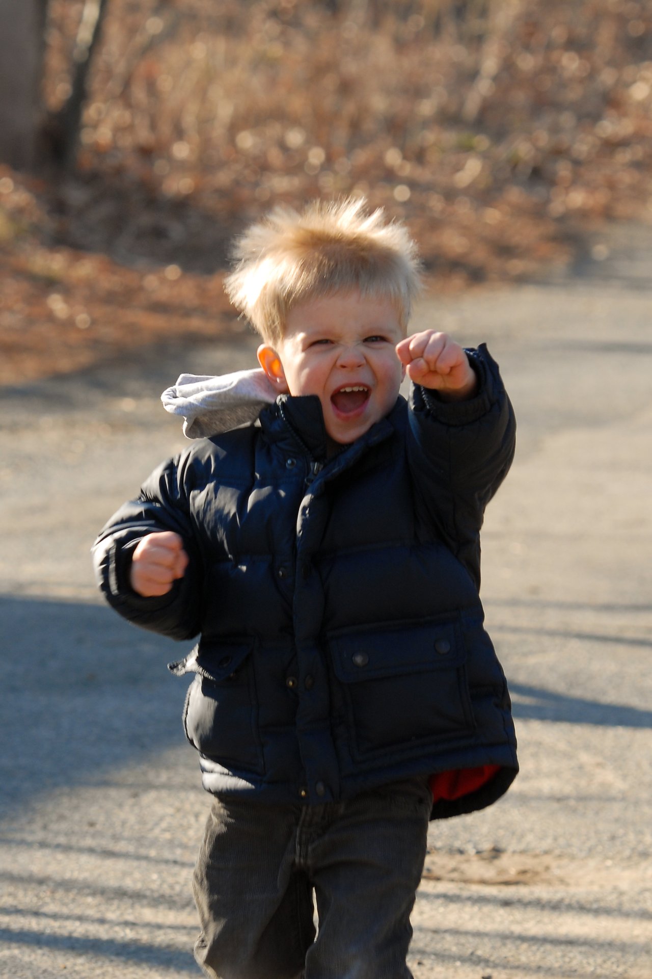A young child in a puffy jacket excitedly raises a fist while walking outdoors on a sunny day.