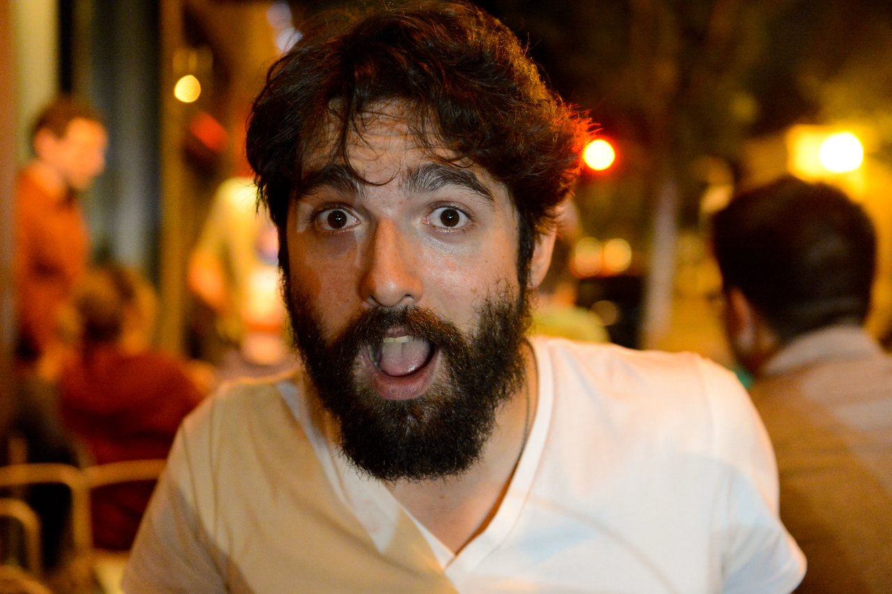 A bearded man in a white shirt looks surprised with his mouth open, sitting in a busy outdoor setting at night.