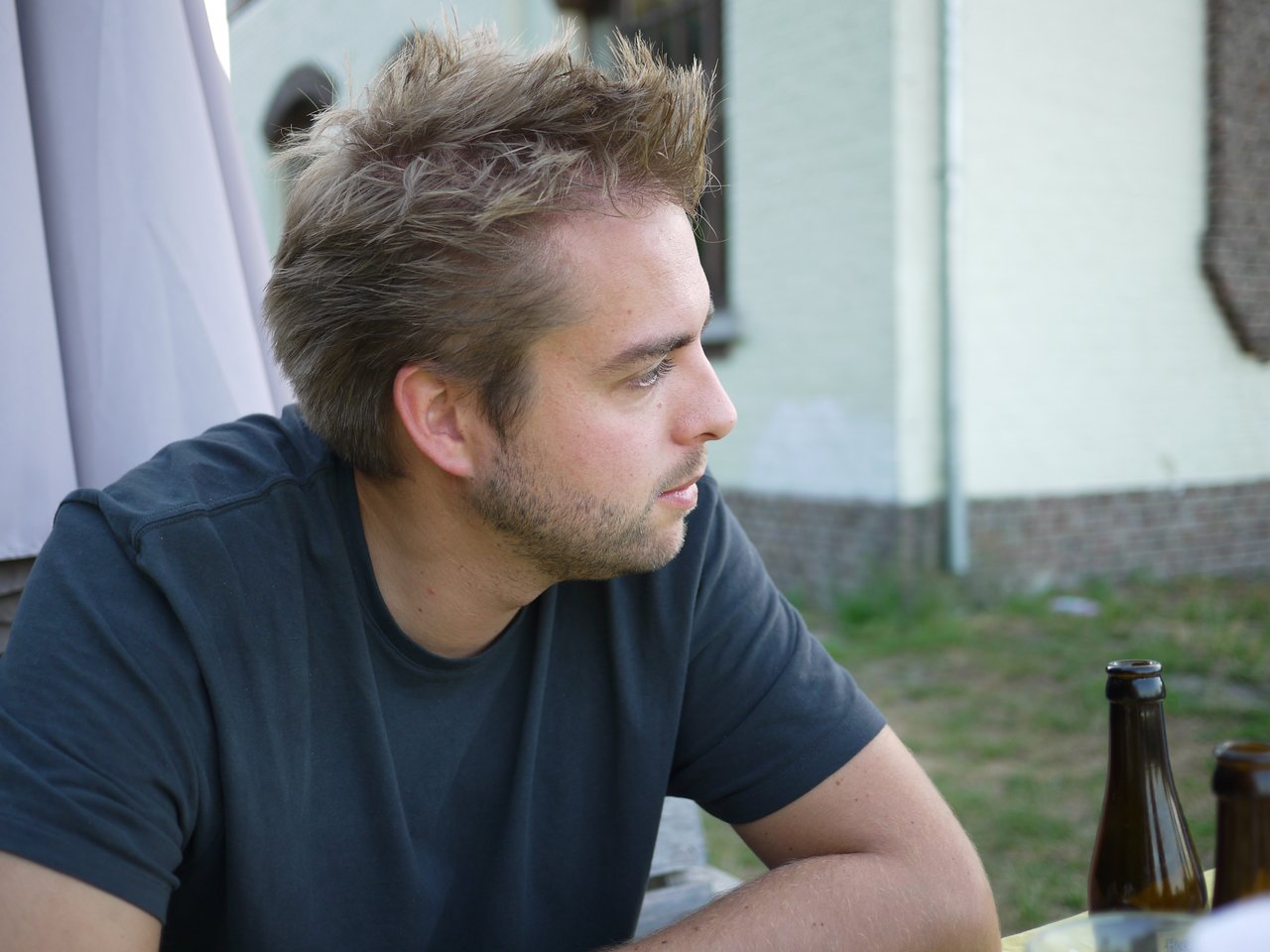 A man in a black shirt sits at an outdoor table, looking to the side with two bottles nearby.