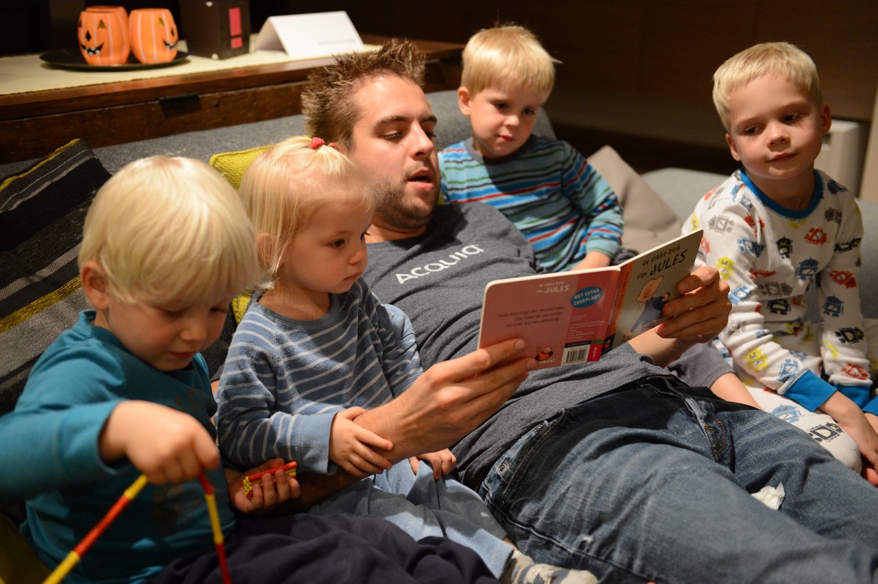 A man reads a bedtime story to four young children sitting closely around him on a couch.
