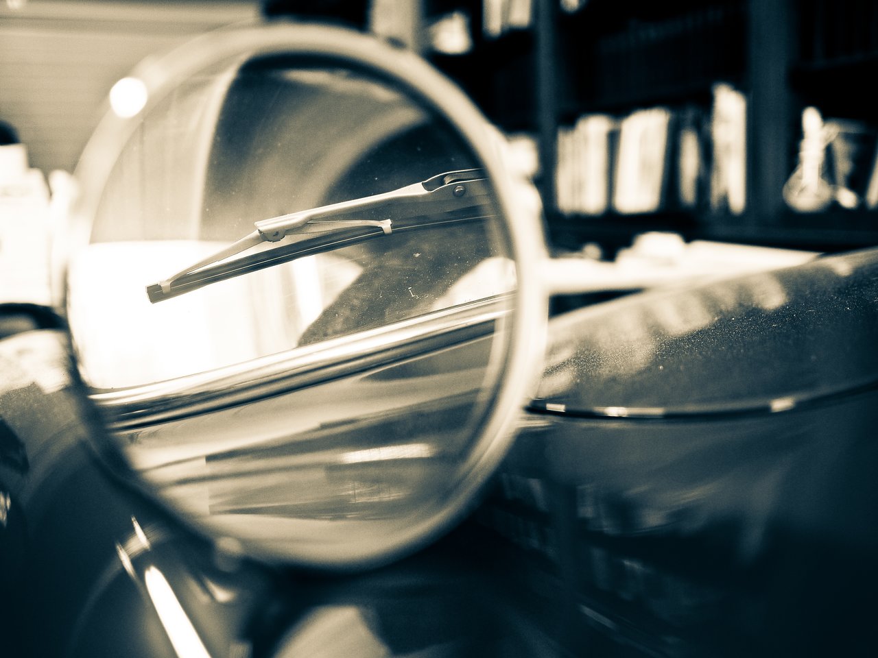 Close-up of a car's side mirror reflecting the windshield and wiper, with a blurred background of shelves.