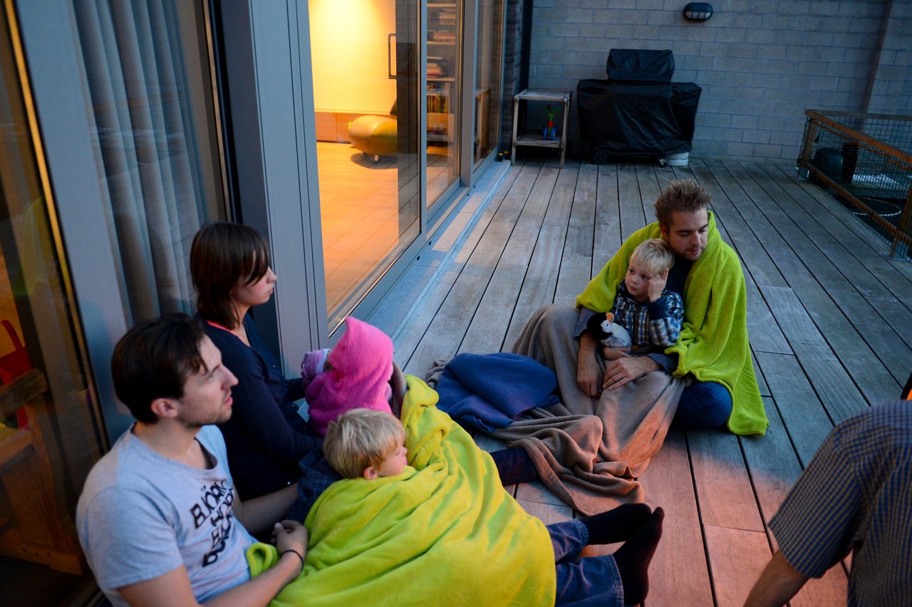 A group of people sit on a wooden deck, wrapped in blankets, gathered around a light source.