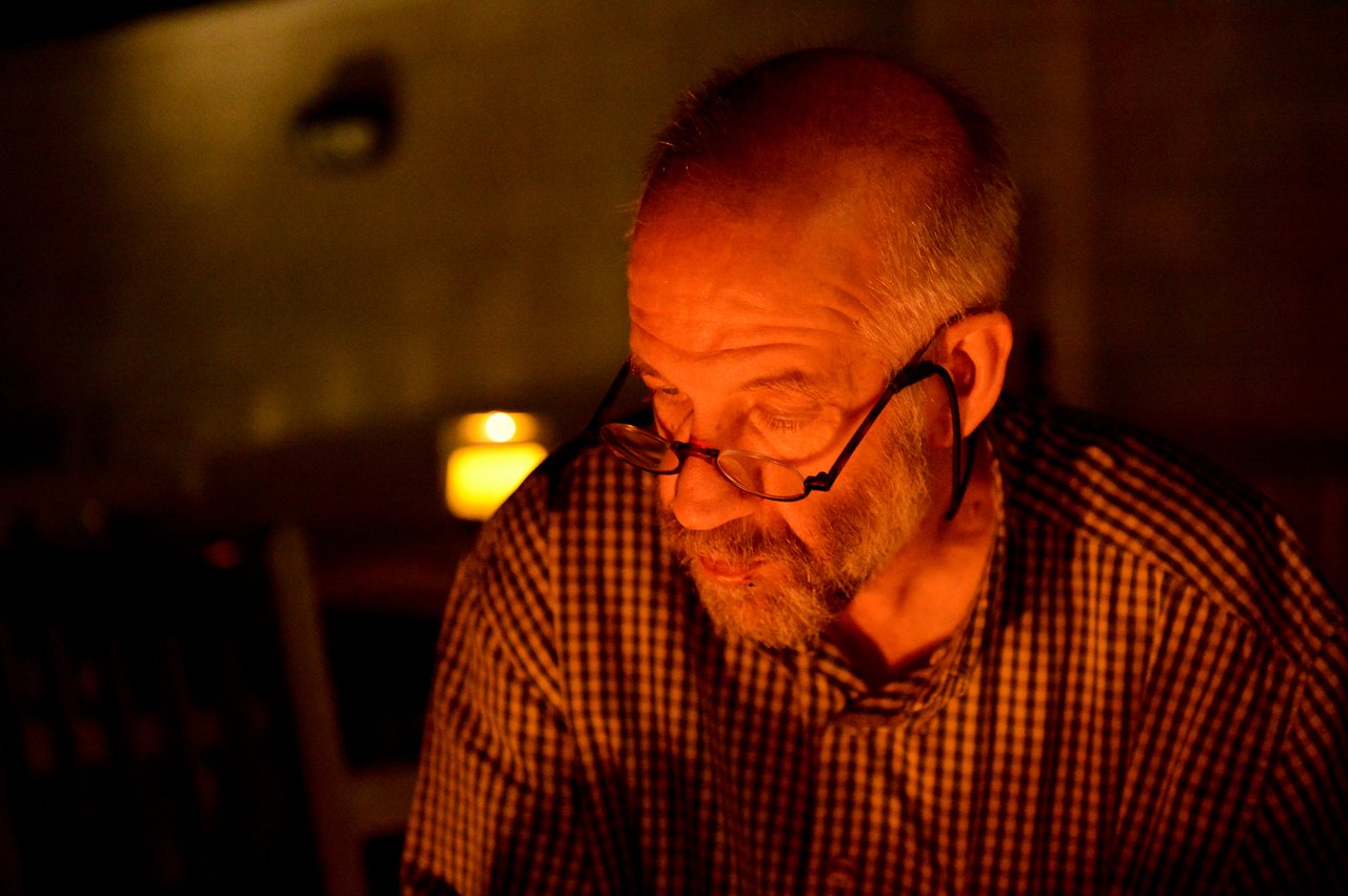 An older man wearing glasses and a checkered shirt looks down, illuminated by warm firelight in a dim setting.