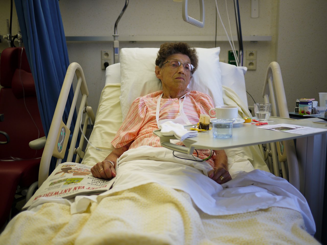Elderly woman lying in a hospital bed with a meal tray and newspaper beside her.