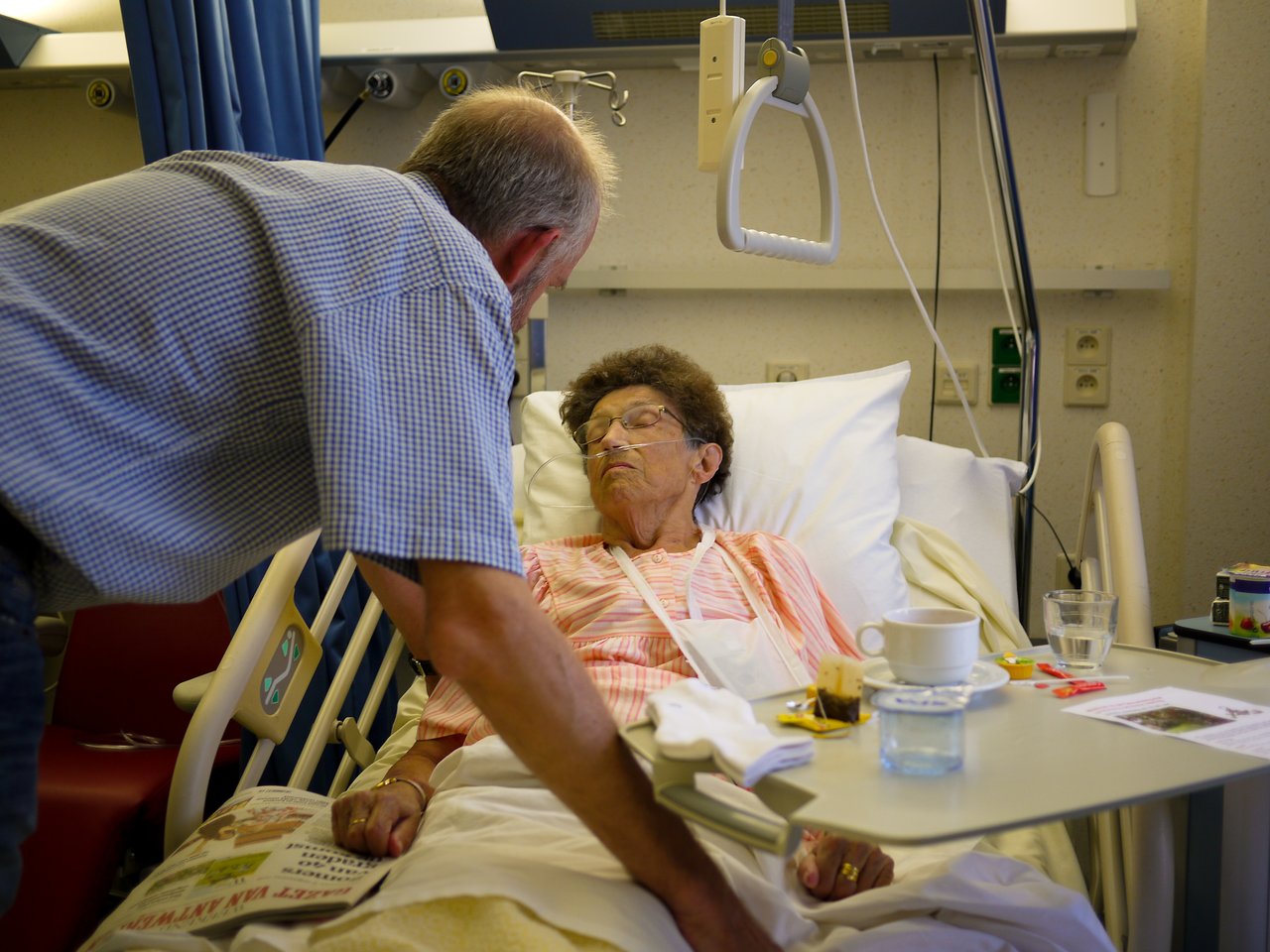 An elderly woman lies in a hospital bed while a man leans in to talk to her.