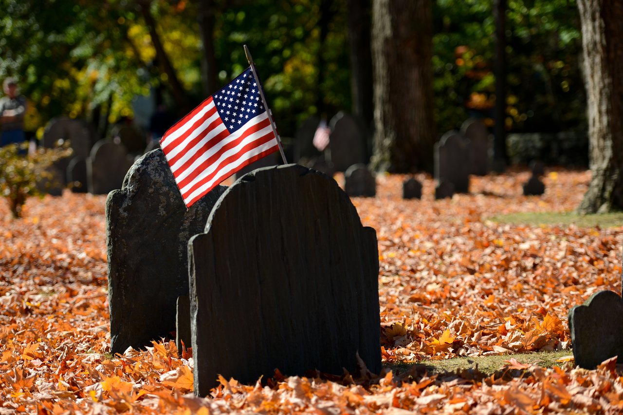 A cemetery with old gravestones covered in fallen leaves, featuring an American flag placed on one of the graves.