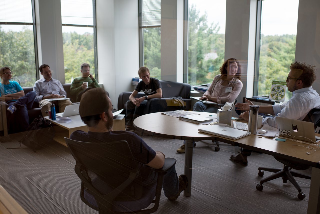 A group of people in an office sit around a table, engaged in a discussion with one person gesturing.