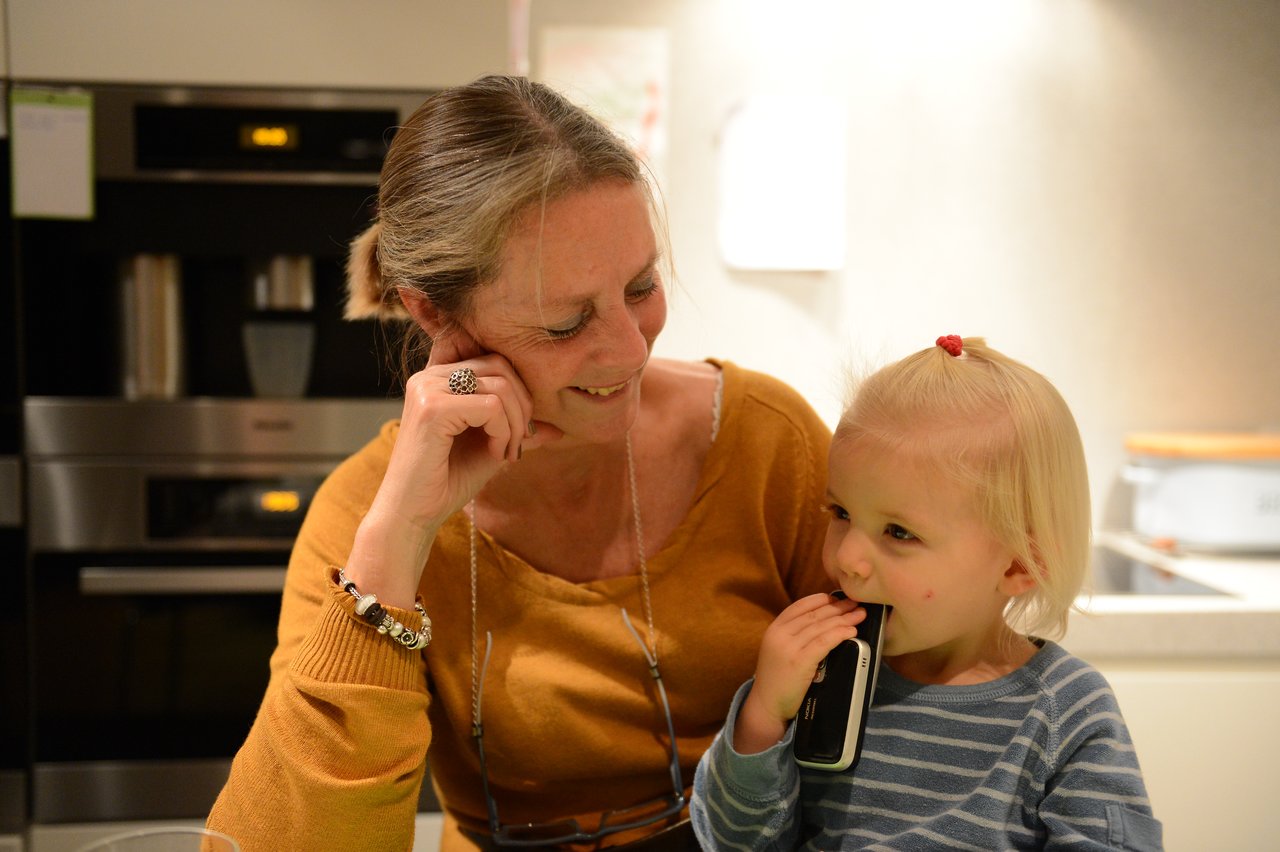 A woman smiles while sitting next to a young child who is holding a phone to their mouth.