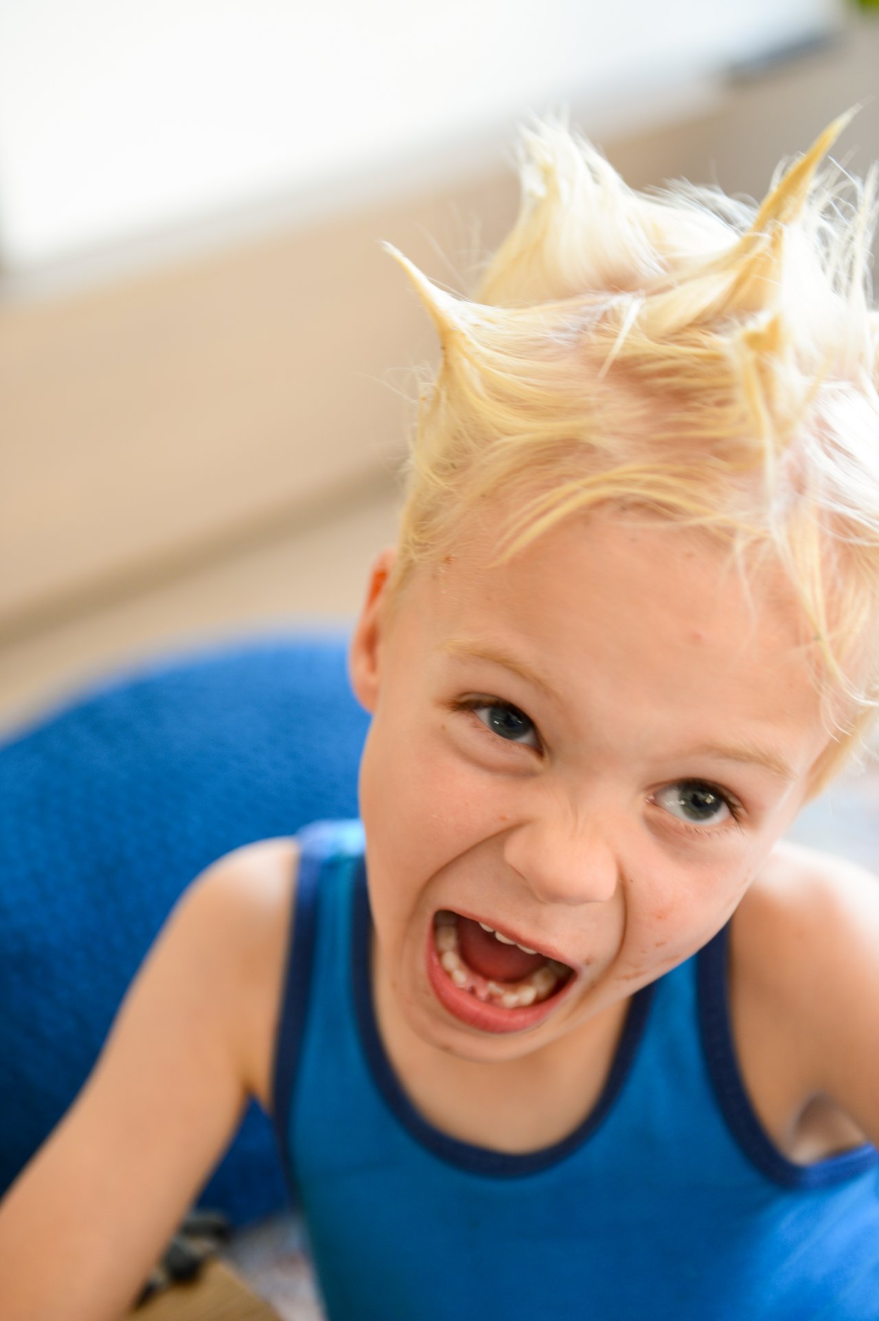 A child with spiky blonde hair and a blue tank top makes a playful, energetic expression while looking forward.