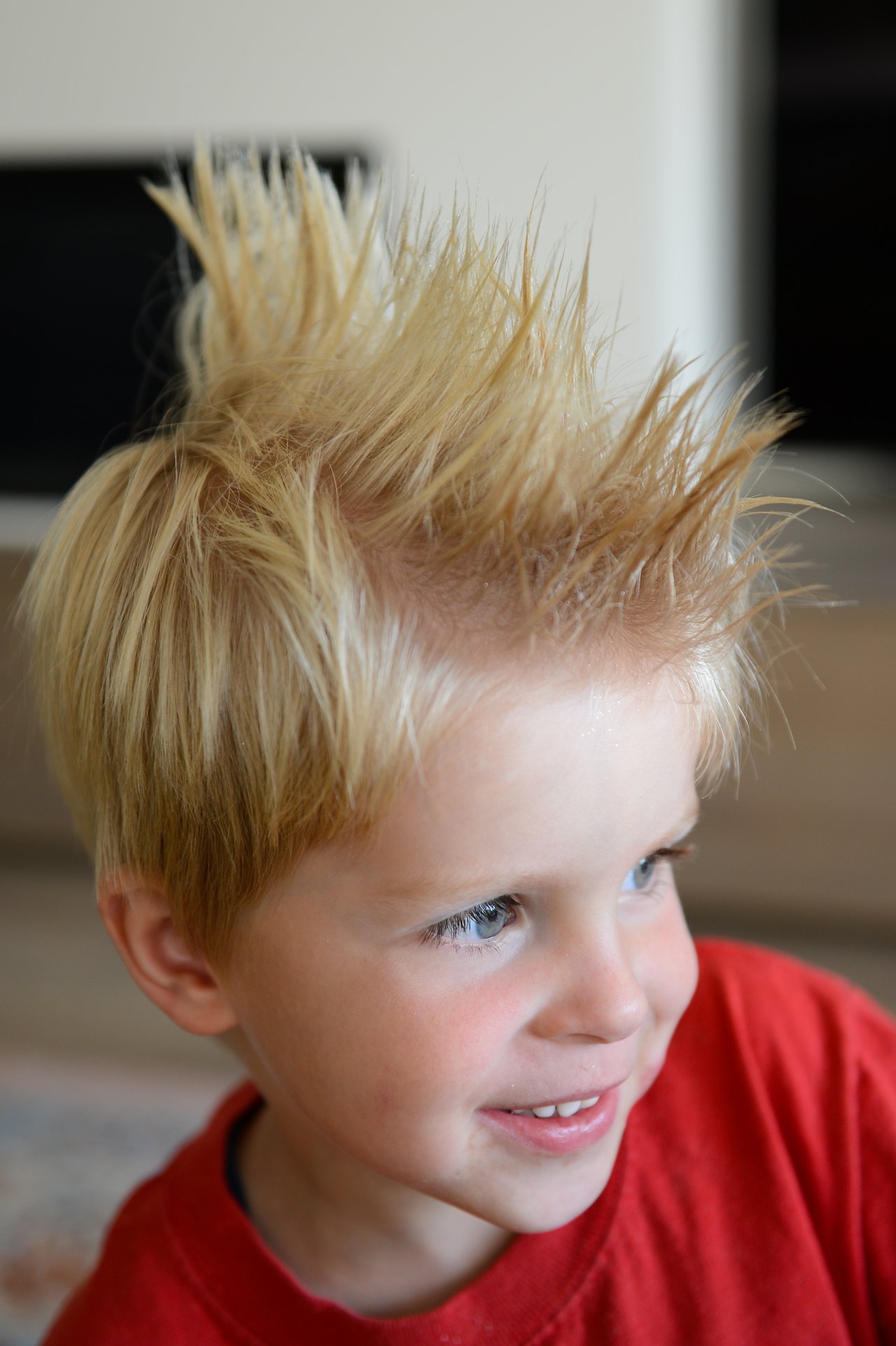 A young child with spiky blonde hair smiles while wearing a red shirt indoors.