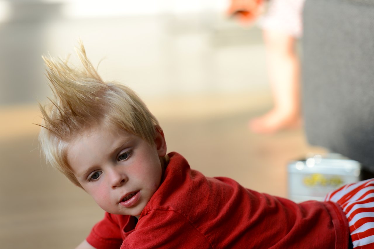 A young child with spiky blonde hair wears a red shirt and leans forward, looking slightly downward.