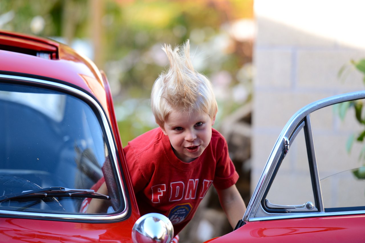 A young child with spiky hair leans out of a red car, smiling playfully while wearing a red shirt.