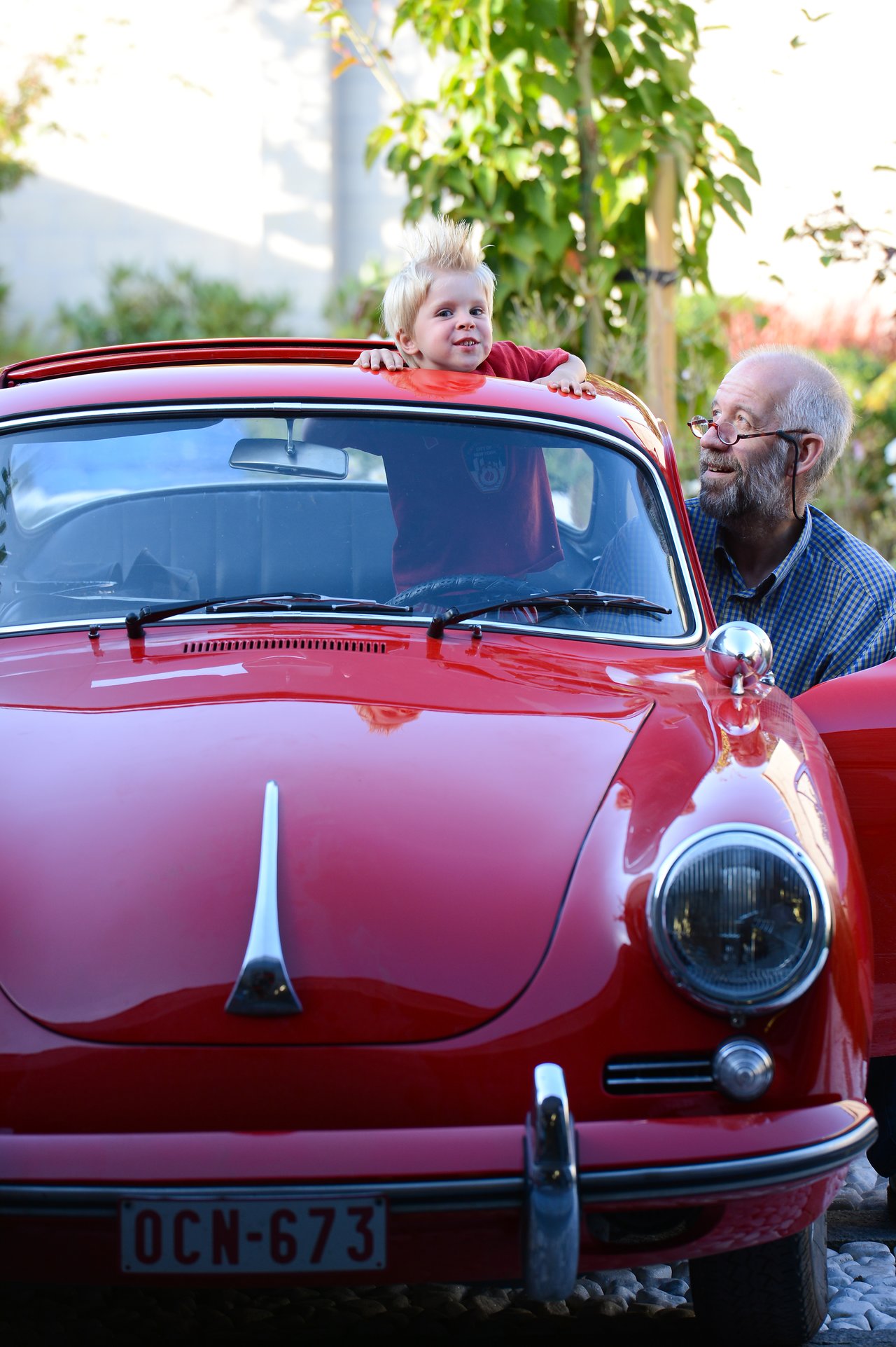 A young child with spiky hair leans out of a red vintage car's sunroof while an older man watches nearby.