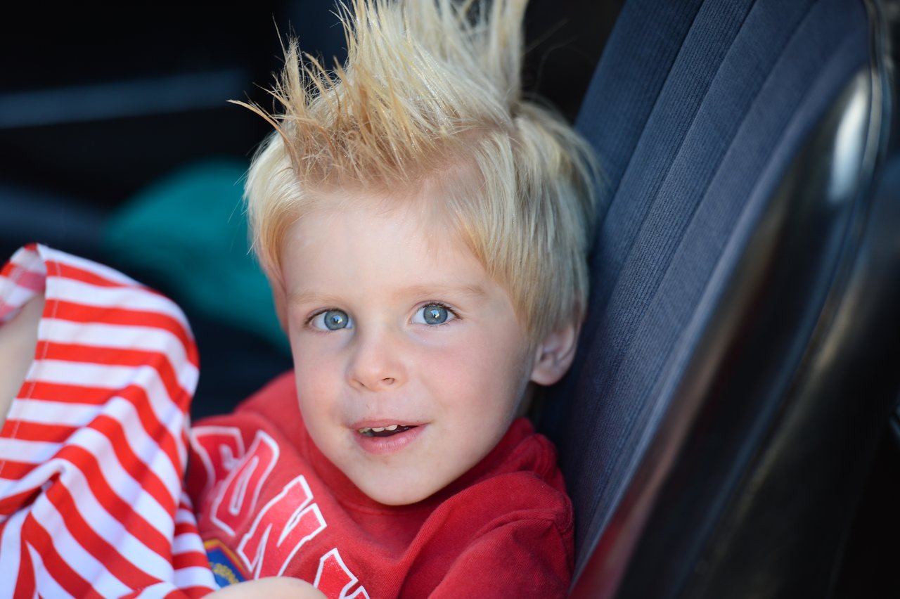 A young child with spiky blonde hair sits in a car seat, wearing a red shirt and smiling slightly.