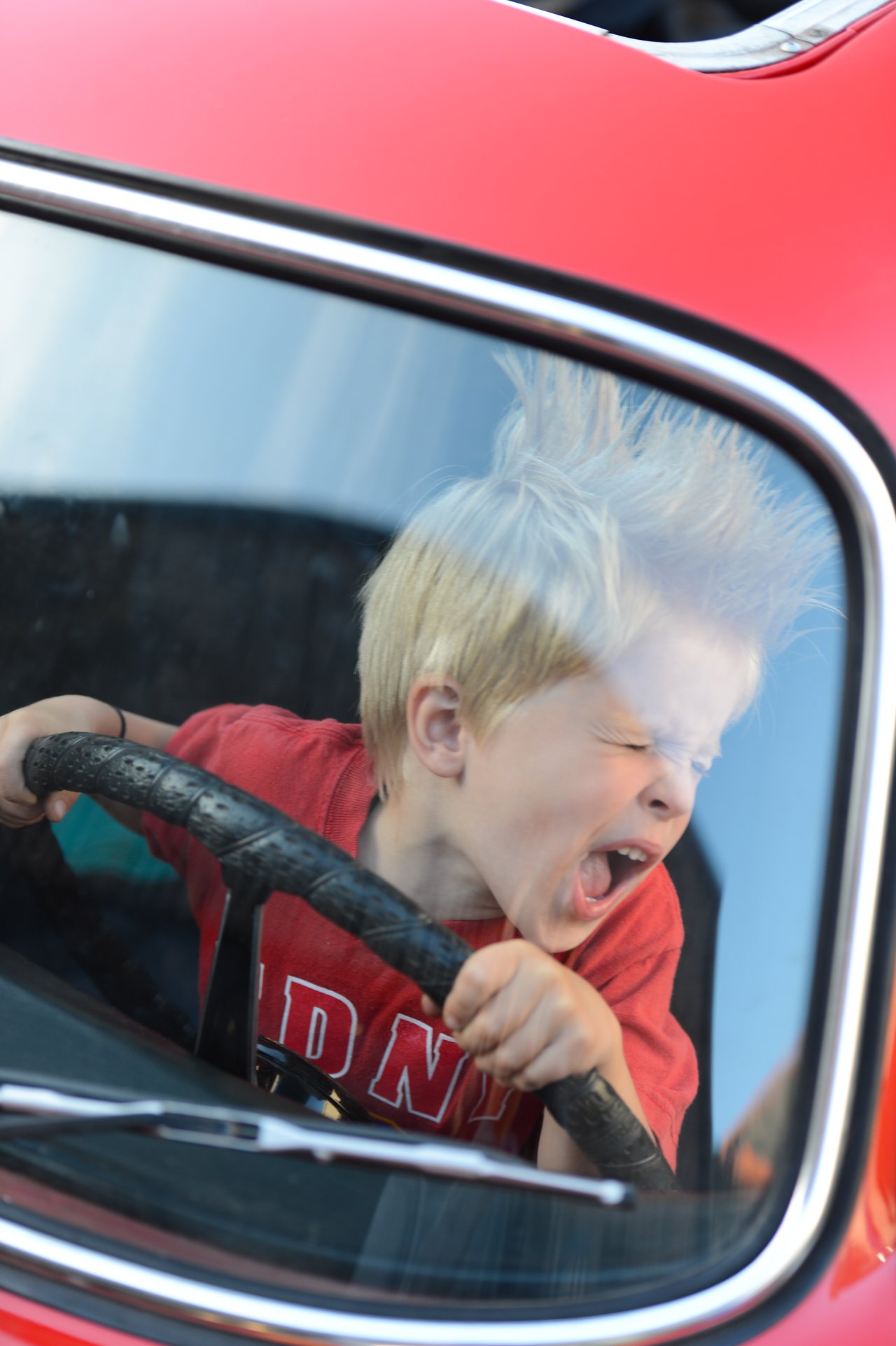 A young child with spiky hair grips a steering wheel and shouts excitedly inside a red car.