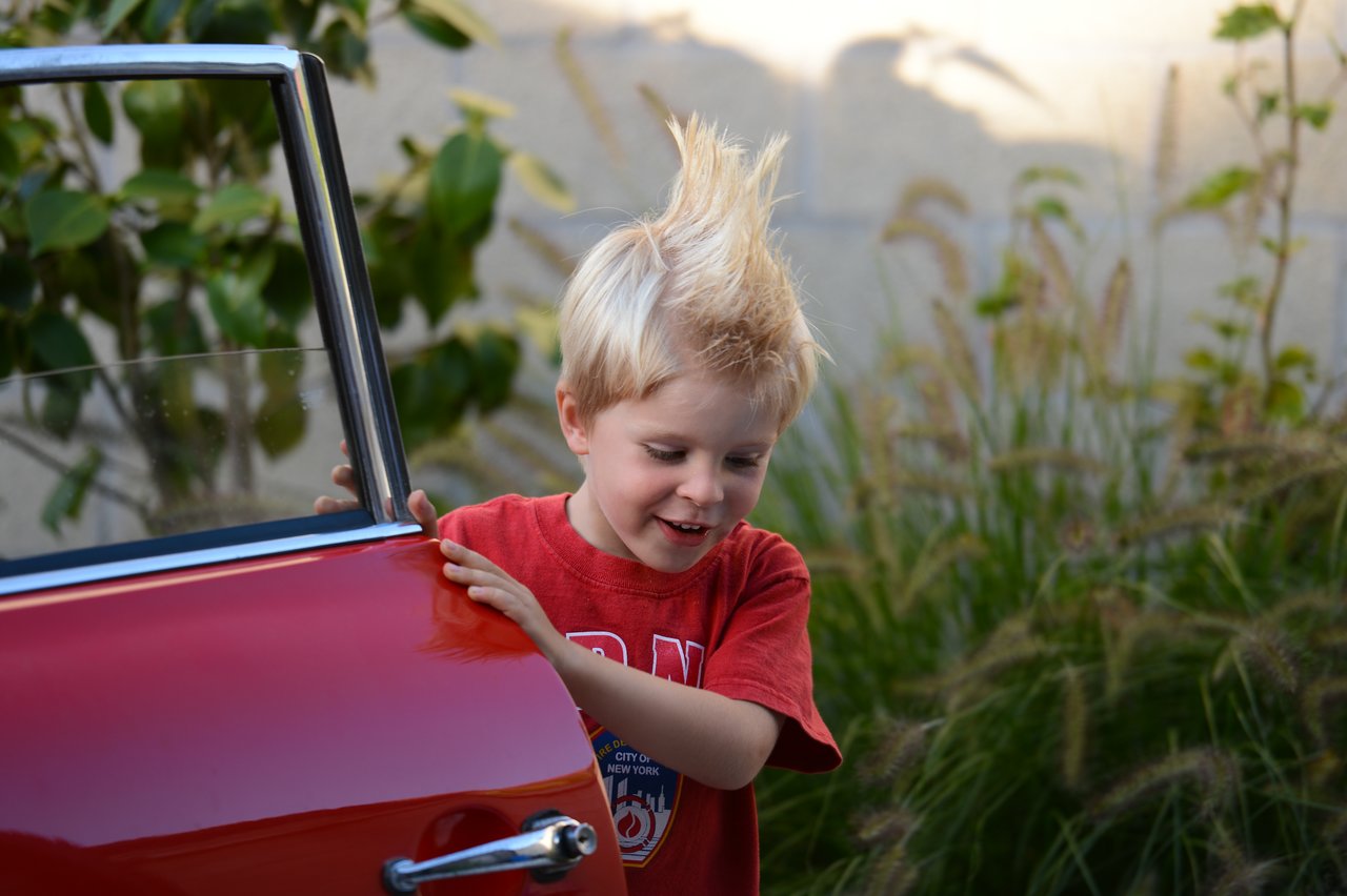 A young child with spiky blond hair holds onto a red car door while smiling and looking down.