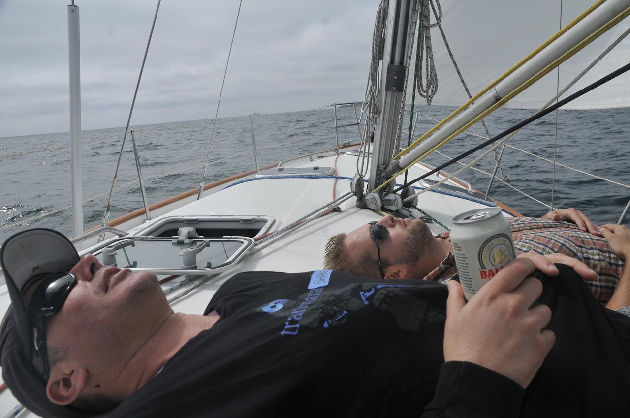 Two people relax on a sailboat, lying down with sunglasses, one holding a can of beer.