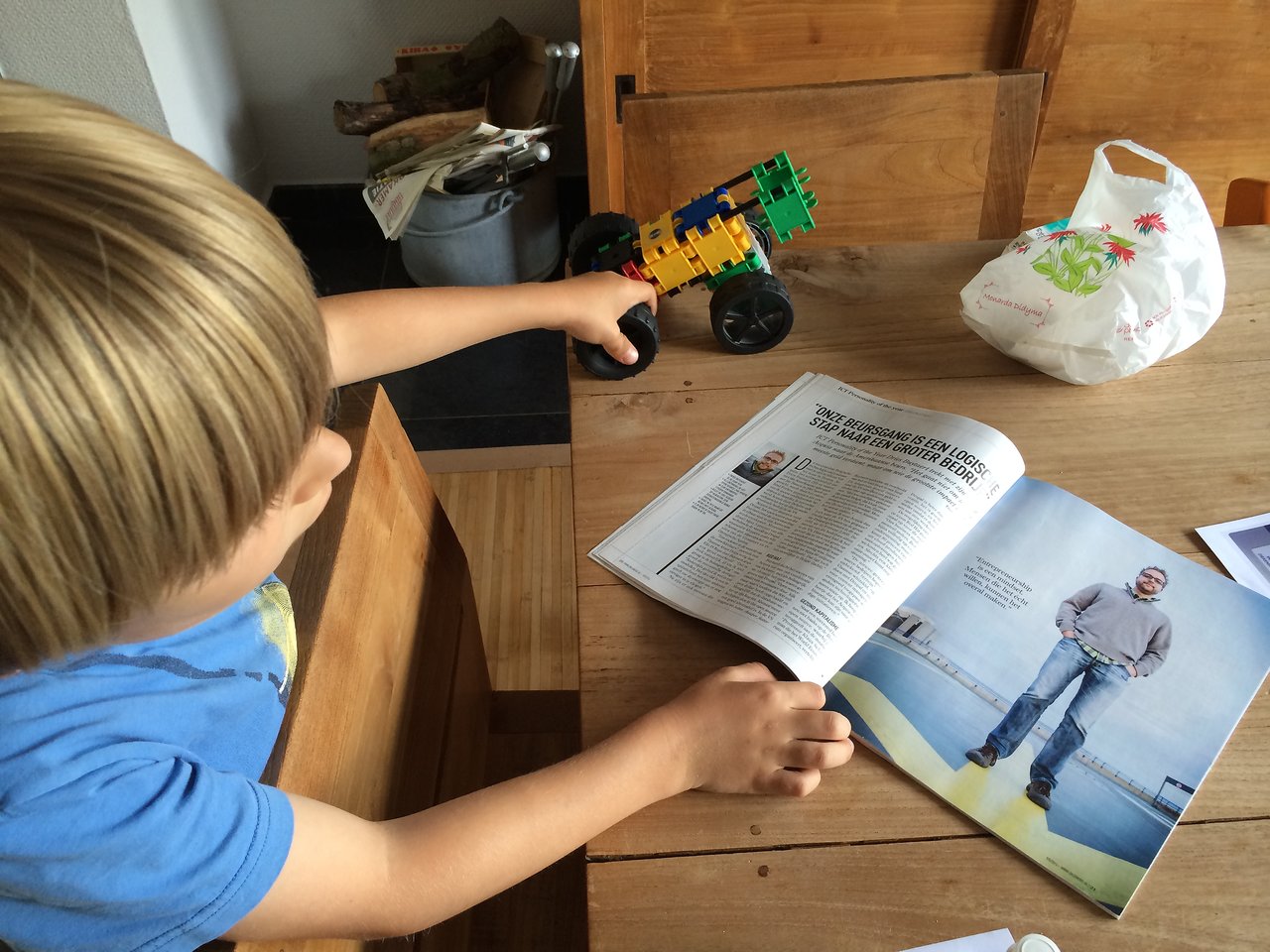 A child in a blue shirt holds a toy car while looking at an open magazine on a wooden table.
