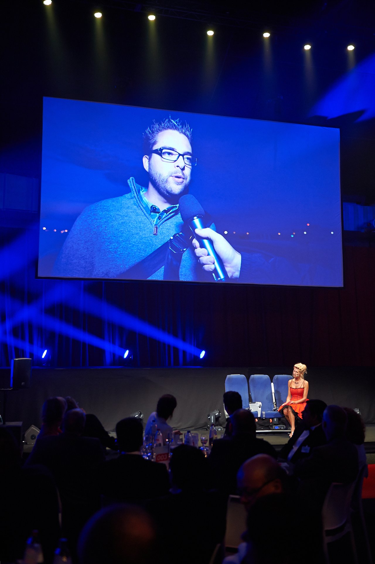 A large screen displays a man being interviewed, while an audience watches and a woman in red sits on stage.