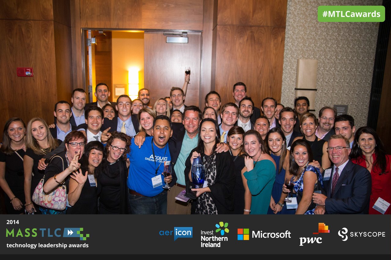 A large group of people smiles and celebrates as a woman in the center holds an award at the MassTLC event.