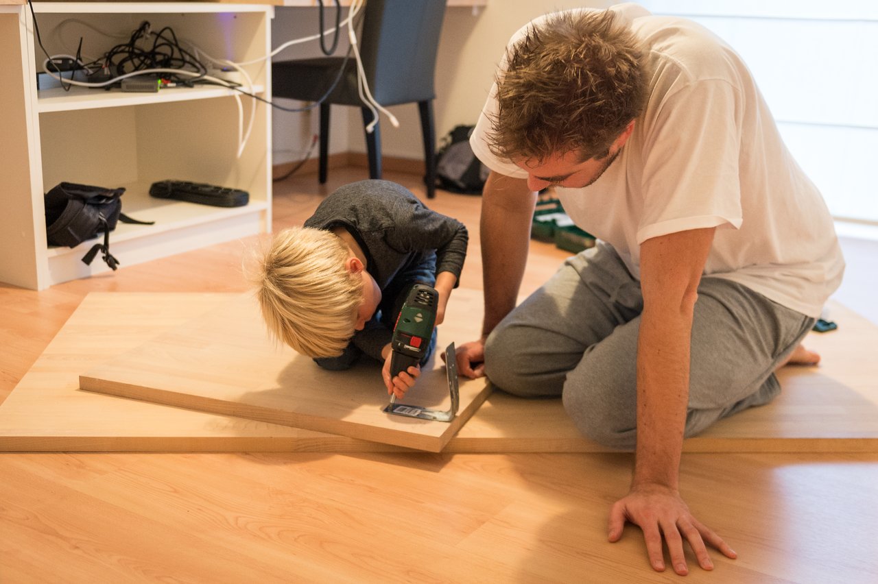 A young child uses a drill while an adult watches closely, both working together on a wooden project.