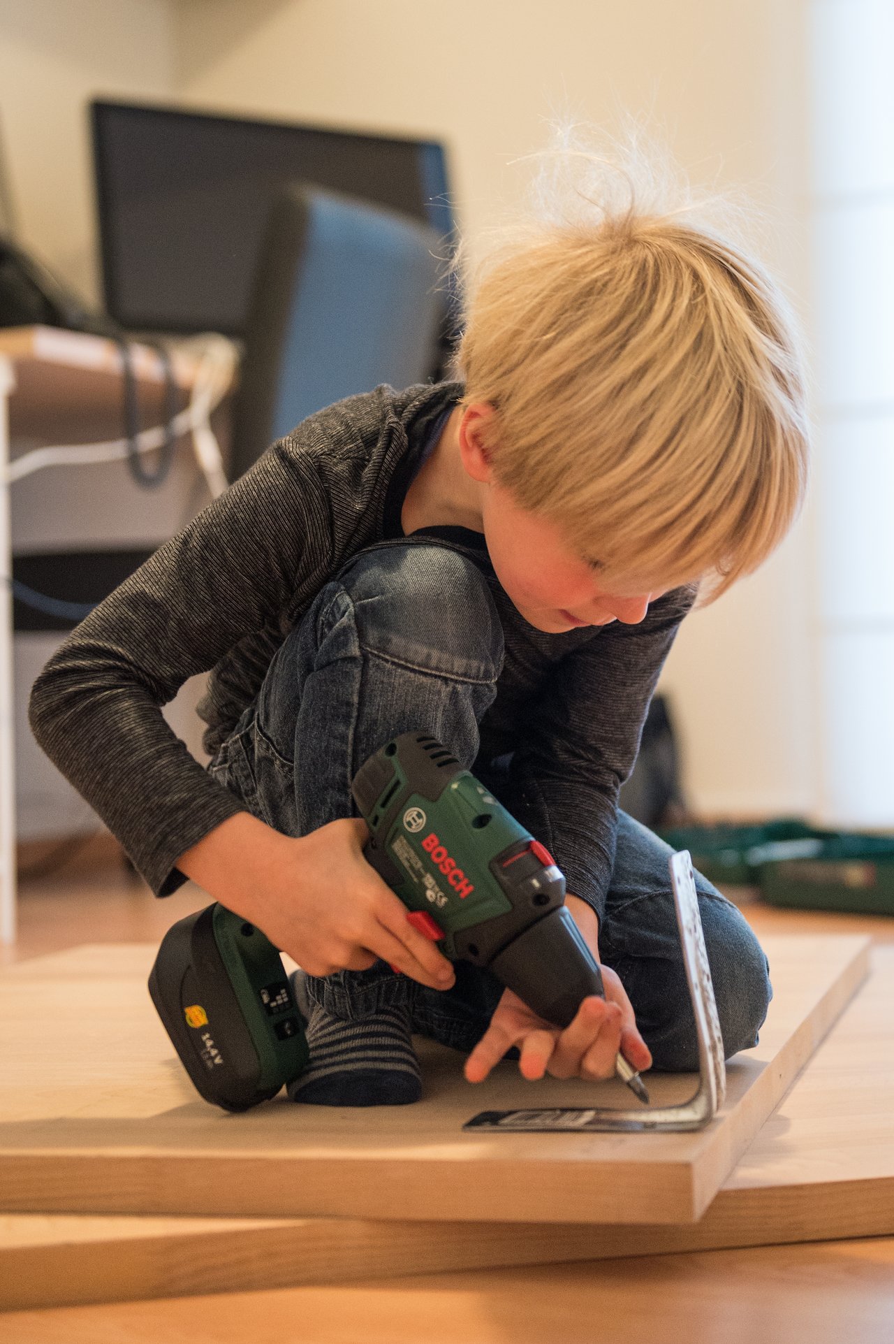 A young child uses a power drill to help with a woodworking project, holding a metal bracket in place.