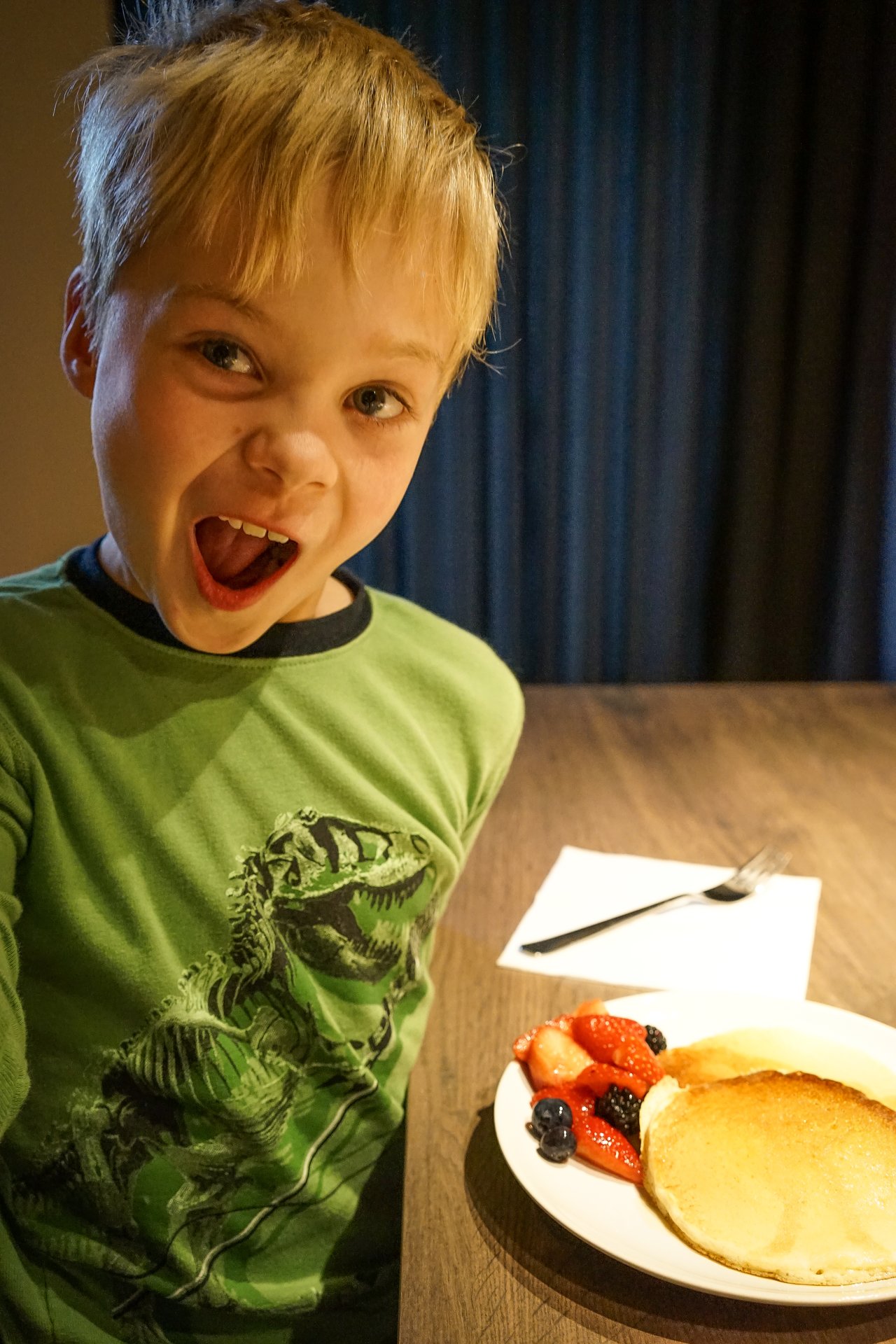 A child in a green dinosaur shirt looks excited while sitting at a table with pancakes and fruit.