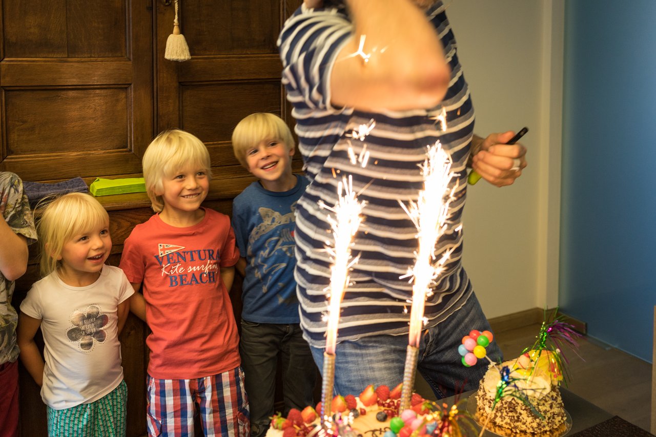 A person's arm is close to sparklers on a cake, with children watching and smiling in the background.