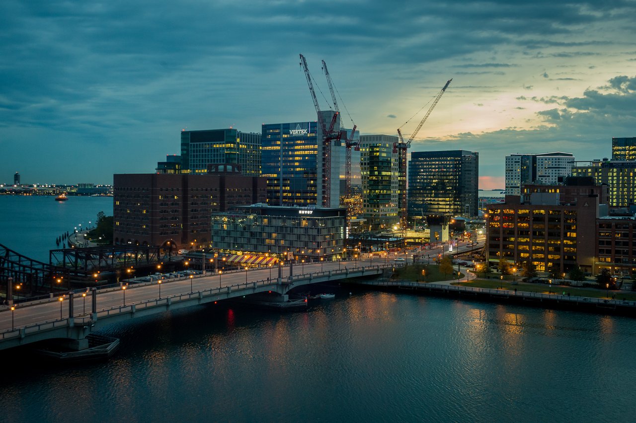 A cityscape of Boston in the early morning, showing buildings, cranes, a bridge, and lights reflecting on water.
