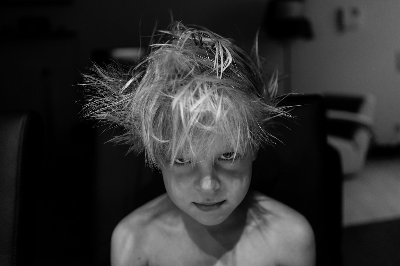 A child with messy, spiky hair sits shirtless, looking directly at the camera with a slight smile.