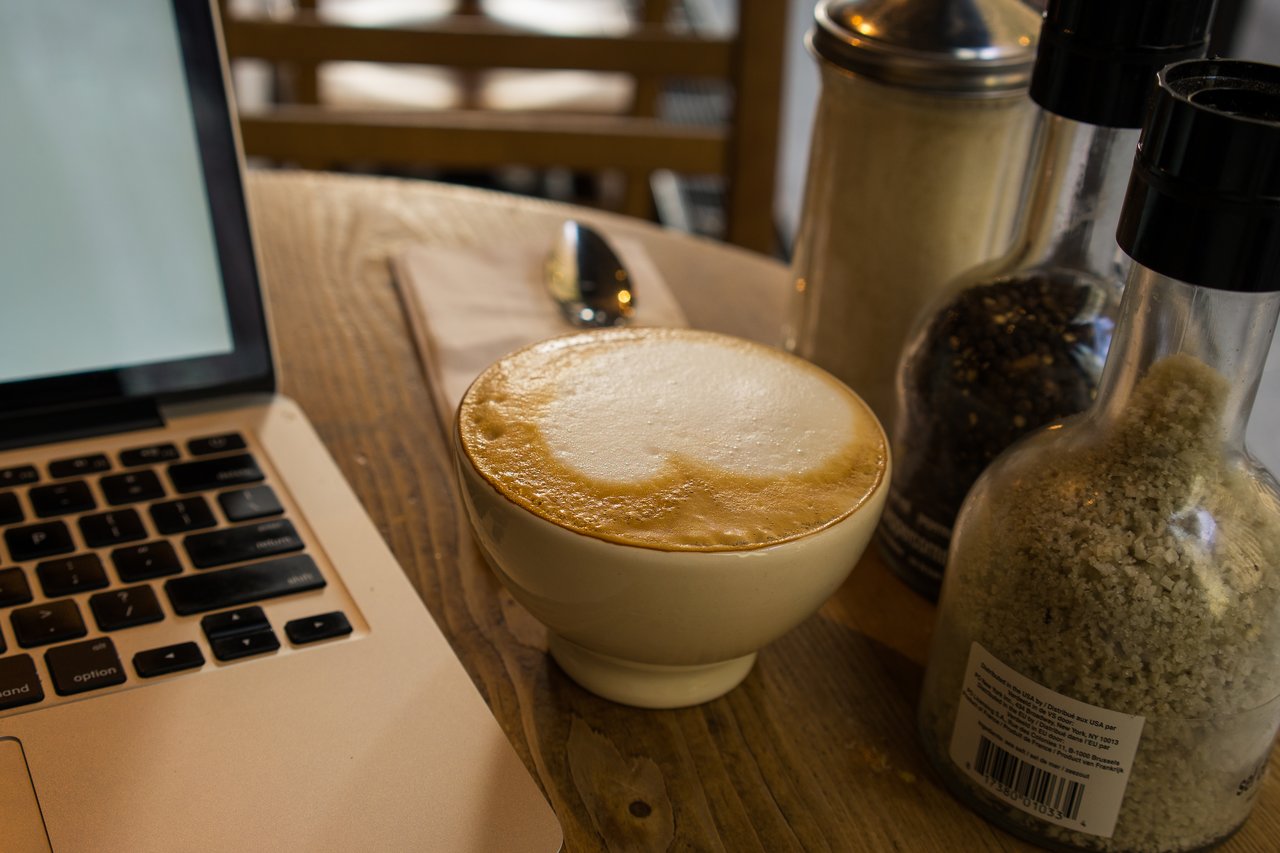 A cup of cappuccino on a wooden table next to a laptop, with condiments and a napkin nearby.